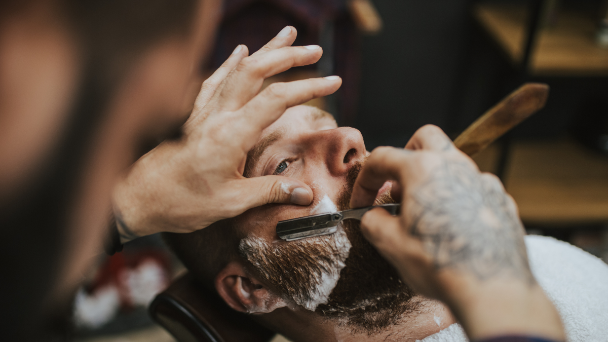 A barber cuts his client's beard while discussing cosmetology licensure.
